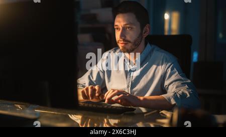 Happy Success Manager travaille sur un ordinateur de bureau dans Creative Agency dans Dark Loft Office dans la soirée. Décoration élégante rénovée avec plantes maison, affiches artistiques et grandes fenêtres arrondies. Banque D'Images