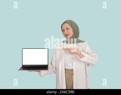 portrait de jeune fille musulmane caucasienne excitée en hijab présentant un ordinateur portable. Tenant un carnet vide à écran vierge d'une main, pointant la maquette vers le haut Banque D'Images