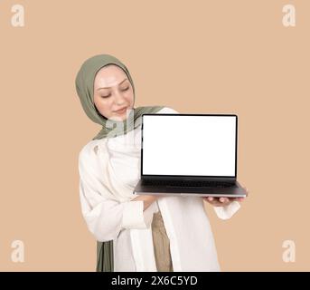 Jeune femme musulmane en hijab tenant une maquette d'ordinateur portable. Fille caucasienne portant un foulard de tête regardant l'écran vide blanc blanc du cahier moderne. Banque D'Images