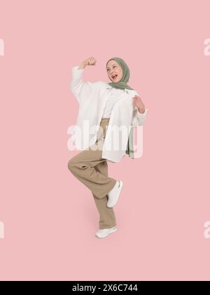 plein corps excité le vainqueur caucasien femme musulmane posant sur fond rose de studio. Portez le hijab et l'abaya en faisant un geste de célébration serrant les poings. Banque D'Images