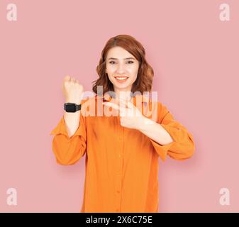 Regarde le temps ! Heureux positif jeune tête rouge bob fille aux cheveux pointant l'index sur la montre intelligente sur son poignet. Appareil photo souriant. Banque D'Images