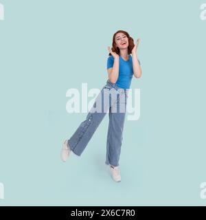 Portrait studio de jeune femme heureuse de corps plein. Debout sur une jambe. Danser, se tenir les mains près de la bouche ouverte. Fond bleu clair isolé. Banque D'Images