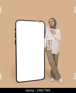 Jeune fille musulmane caucasienne pleine de corps en hijab debout près et pointant du doigt vers une énorme maquette de téléphone portable. Vider l'écran blanc vierge. Banque D'Images