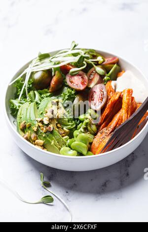 Saladier végétalien avec patate douce cuite au four, haricots edamame, tomates, avocat, noix, dressing et pousses de tahini, fond de marbre blanc. Banque D'Images