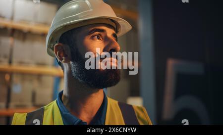 Portrait d'un travailleur latin portant un casque de sécurité debout à l'extérieur de l'entrepôt souriant. Héros professionnel de première ligne portant une barbe élégante debout dans une lumière cinématographique Banque D'Images