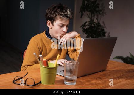 Adolescent étudiant à la maison avec un ordinateur portable, concentré et productif, entouré de matériel d'étude, dans un environnement domestique confortable. Banque D'Images