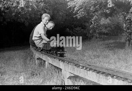 années 1960, historique, à l'extérieur dans une zone herbeuse d'un grand jardin, un jeune homme avec son père à cheval sur un train à vapeur miniature sur une voie ferrée en béton surélevée. Banque D'Images