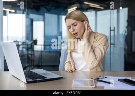 Une femme d'affaires professionnelle éprouvant de graves douleurs à l'oreille, peut-être une otite, tout en travaillant dans un cadre de bureau contemporain. Banque D'Images