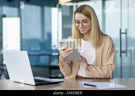 Une femme professionnelle souriante dans un blazer beige utilise une tablette assise à une table en verre dans un cadre de bureau contemporain, avec un ordinateur portable et des documents. Banque D'Images