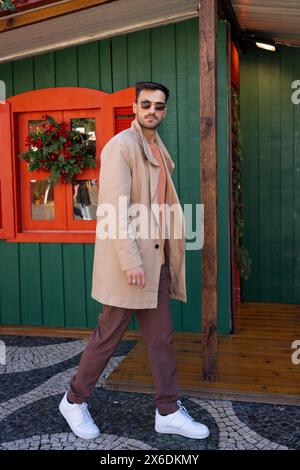 Corps entier de bel homme confiant dans des lunettes de soleil et trench trench beige à la mode marchant à l'extérieur de la maison décorée de Noël. Vue latérale du mâle hipster dans la tenue à la mode en ville. Banque D'Images