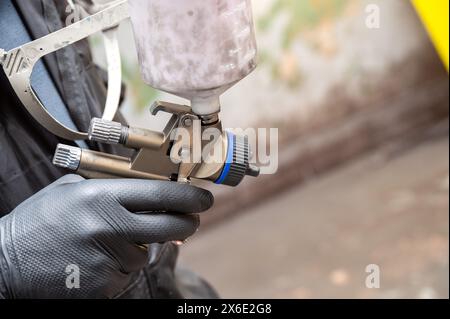 Gros plan de la main d'un peintre professionnel tenant un pistolet de pulvérisation avec un récipient de peinture, prêt pour le travail de carrosserie automobile Banque D'Images