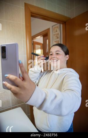 Une jeune femme se maquille à la maison, améliorant sa beauté naturelle. Elle applique fond de teint, correcteur, blush, fard à paupières, eyeliner, mascara et lipstique Banque D'Images