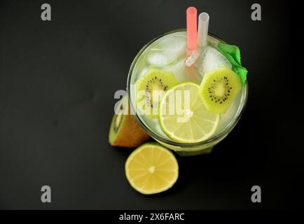 Un verre d'un mélange de jus de fruits avec de la glace et des pailles sur un fond noir, à côté de kiwi mûrs et de citron vert. Vue de dessus, pose à plat. Banque D'Images