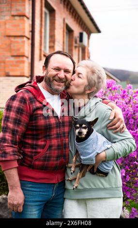 Couple marié russe mature embrassant et serrant près du buisson fleuri de rhododendron, tenant un chien Banque D'Images