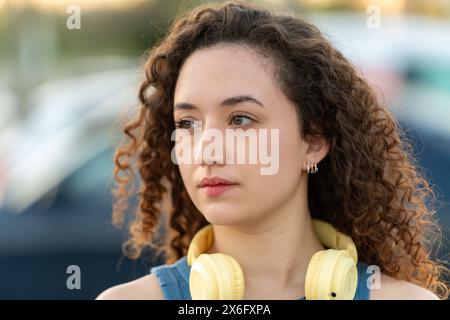 Portrait d'une jeune femme réfléchie avec des cheveux bouclés et des écouteurs autour du cou, regardant contemplatif dans un cadre urbain extérieur au crépuscule. Banque D'Images