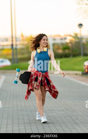 Jeune femme joyeuse tenant une planche à roulettes et portant des écouteurs, marchant décontractée dans un parc ensoleillé, avec un débardeur bleu et une chemise à carreaux. Banque D'Images
