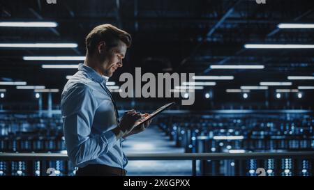 Expert INFORMATIQUE souriant utilisant un ordinateur tablette dans un centre de données. Homme d'affaires prospère et entrepreneur en e-business surplombant l'installation de Cloud Computing de ferme de serveurs. Banque D'Images