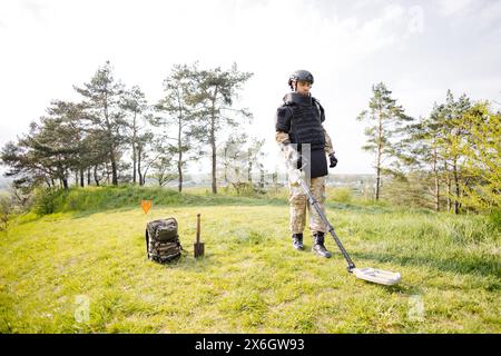 Un homme dans un uniforme militaire et gilet à l'épreuve des balles travaille dans la forêt avec un détecteur de métaux. Un mineur effectue des travaux de déminage du territoire. Banque D'Images