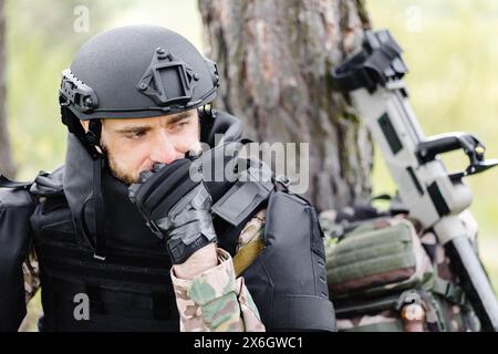 Un homme dans un uniforme militaire et un gilet à l'épreuve des balles se trouve dans la forêt près d'un détecteur de métal et d'un sac à dos militaire. Un homme s'arrête dans le travail de demin Banque D'Images