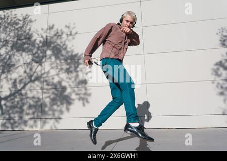 Jeune homme latin avec des cheveux blonds courts portant des vêtements de style rétro, un pantalon et une chemise marron, à l'extérieur, il est en saut d'air avec des écouteurs écoutant Banque D'Images