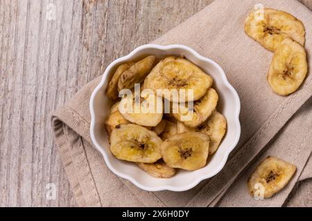 Vue de dessus, extrême gros plan, sur un fond en bois texturé, un bol avec des chips de banane déshydratées Banque D'Images