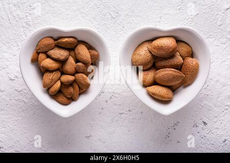 Vue de dessus, gros plan extrême, sur fond de béton blanc texturé, deux bols en forme de cœur avec des amandes dans la coquille et sans. Banque D'Images