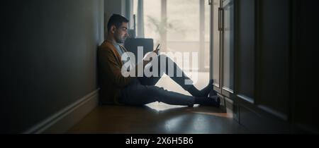 Portrait de jeune homme adulte utilisant smartphone à la maison. Il est assis au sol du couloir, dos au mur. Utilise Internet pour consulter le Web et discuter, divertissement sur les médias sociaux. Banque D'Images