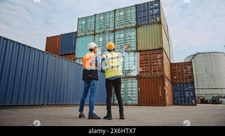 Bel ingénieur industriel et superviseur afro-américain en casques de sécurité et gilets de sécurité debout dos à la caméra dans le terminal à conteneurs. Les employés parlent des opérations logistiques. Banque D'Images