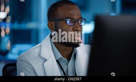 Portrait de bel homme noir portant des lunettes travaillant en toute confiance sur un ordinateur. Jeune ingénieur ou scientifique masculin intelligent travaillant en laboratoire. Fond bleu bokeh. Gros plan Banque D'Images