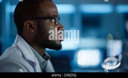 Portrait de bel homme noir portant des lunettes travaillant en toute confiance sur un ordinateur. Jeune ingénieur ou scientifique masculin intelligent travaillant en laboratoire. Fond bleu bokeh. Gros plan vue latérale Banque D'Images