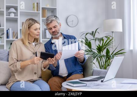 Couple mature assis sur un canapé, examinant des documents et utilisant une tablette à la maison. Ils semblent concentrés et engagés dans une discussion financière. Banque D'Images
