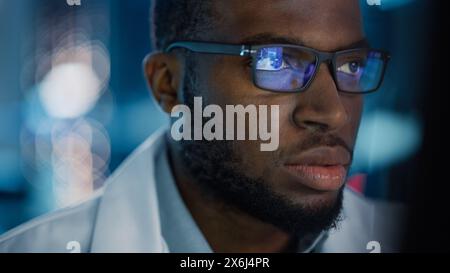 Portrait de bel homme noir portant des lunettes travaillant en toute confiance sur un ordinateur. Jeune ingénieur ou scientifique masculin intelligent travaillant en laboratoire. Fond bleu bokeh. Gros plan Banque D'Images
