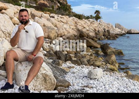 L'image capture un moment de tranquillité d'un homme de 30 ans, assis sur la rive rocheuse, regardant la mer calme sous un ciel clair. Banque D'Images