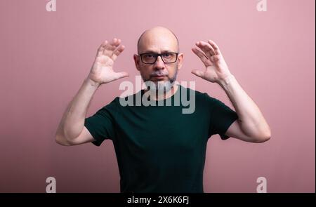 Portrait d'homme chauve et barbu avec des lunettes avec les deux mains à côté de sa tête regardant la caméra. Isolé sur fond de couleur saumon. Banque D'Images