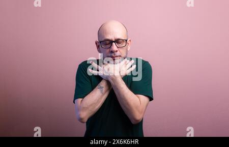 Homme chauve barbu avec des lunettes de vue serrant son cou avec les deux mains. Isolé sur fond de couleur saumon. Banque D'Images
