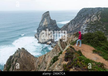 Un randonneur solitaire en veste rouge se tient au bord d'une falaise, surplombant les puissantes vagues de l'océan, immergé dans la grandeur du paysage marin. Un voyage au Portugal le long de la côte de l'océan Atlantique. Banque D'Images