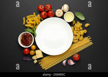Assiette entourée de différents types de pâtes et de produits sur fond noir, pose plate. Espace pour le texte Banque D'Images