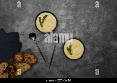 Sur un fond gris, des bols de soupe crémeuse spargel, ornés d’une asperge verte, sont flanqués de toasts de pain, d’élégantes cuillères Banque D'Images
