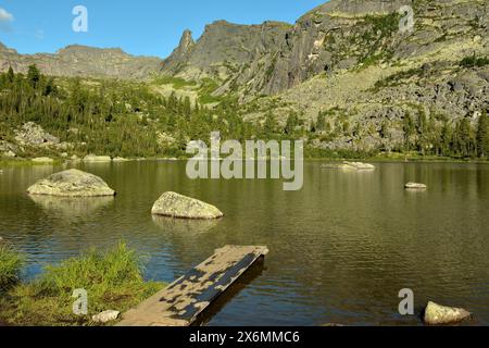 Une plate-forme en bois sort de la rive du lac avec des rebords en pierre à la surface, entourée de hautes montagnes par une journée d'été ensoleillée. Arc-en-ciel l Banque D'Images