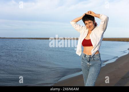 femme aux cheveux courts souriant sur la plage, portant une chemise blanche, un haut rouge et un denim, un ciel bleu et une mer calme. Banque D'Images
