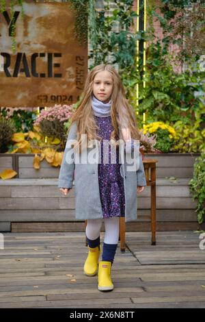 Petite fille en manteau, écharpe et bottes en caoutchouc marchant sur la terrasse Banque D'Images