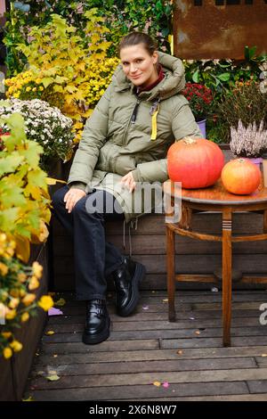 Femme souriante en veste duvet se trouve sur le banc entouré de fleurs d'automne Banque D'Images