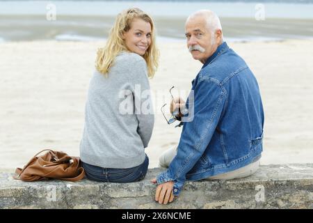 homme senior et jeune femme assis sur le mur surplombant la plage Banque D'Images