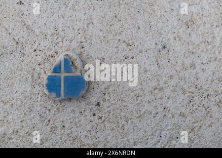 Petite croix gravée en afragment de tuile bleue brisée sur un fond texturé blanc. Banque D'Images