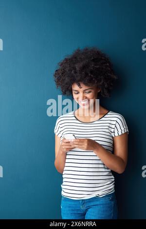 Femme africaine, téléphone et SMS en studio avec sourire, penser et taper sur le web pour le contact par fond bleu. Personne, fille et smartphone dans Banque D'Images