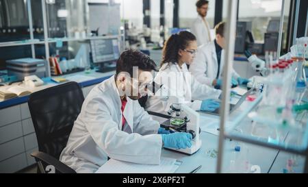 Laboratoire des sciences médicales : rangée de l'équipe diversifiée de jeunes scientifiques multiethniques regardant sous microscope, analyser les produits chimiques, parler, résoudre des problèmes. Spécialistes en biotechnologie travaillant dans Advanced Lab Banque D'Images