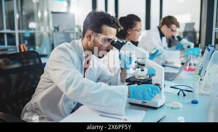 Laboratoire des sciences médicales : rangée de l'équipe diversifiée de jeunes scientifiques multiethniques regardant sous microscope, analyser les produits chimiques, parler, résoudre des problèmes. Spécialistes en biotechnologie travaillant dans Advanced Lab Banque D'Images