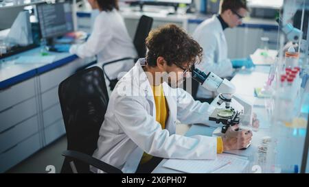 Laboratoire des sciences médicales : beau scientifique latin cherchant sous microscope pour analyser de nouveaux échantillons. Jeune spécialiste en biotechnologie, utilisant des équipements avancés. Banque D'Images