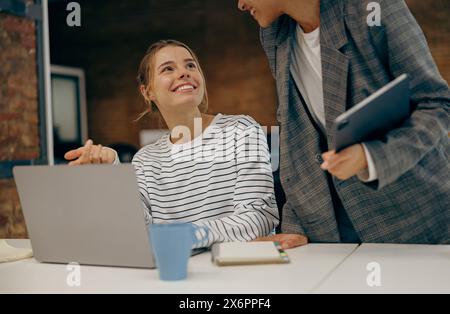 Deux collègues féminines souriantes et occupées travaillant sur un projet ensemble et utilisant un ordinateur portable assis dans le bureau Banque D'Images