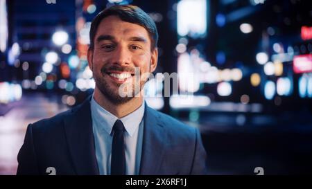 Portrait de bel homme barbu souriant, regardant la caméra, debout dans la ville de nuit avec Bokeh Neon Street Lights en arrière-plan. Heureux confiant jeune homme portant un costume élégant. Banque D'Images